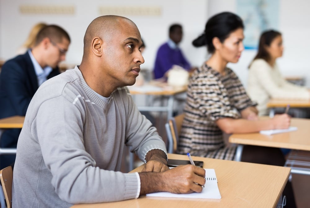 Attentive adult students in a classroom setting, seated at desks and taking notes, with a man in a beige sweater in focus.